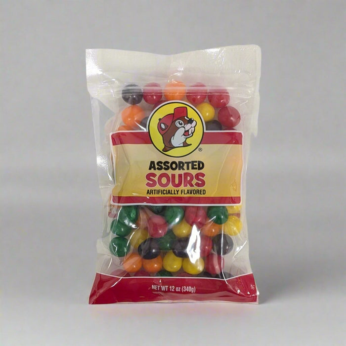 A clear plastic bag of "Assorted Sours" candy is displayed on a white surface. The bag has a red and yellow label with an illustrated character resembling a beaver wearing a hat. The candies inside are small, round, and brightly colored in shades of red, green, yellow, orange, and purple. The packaging indicates a net weight of 12 ounces (340 grams).