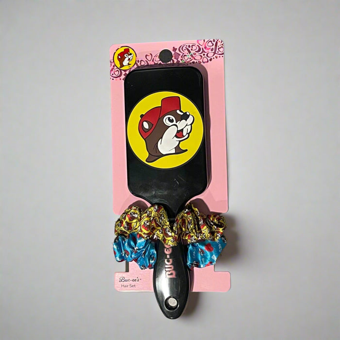 Two Buc-ee’s hair brush and scrunchie sets on display: one with a purple brush and heart scrunchies, the other with a black brush and colorful red-cap Buc-ee's theme scrunchies.