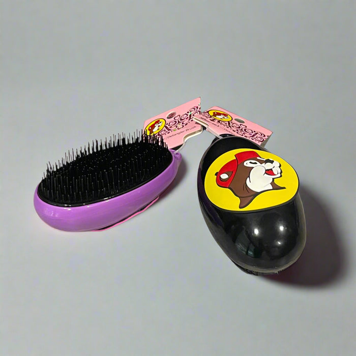 Buc-ee’s detangler brushes in two colors: one black with Buc-ee in a red cap, and one purple with Buc-ee surrounded by pink hearts, shown from front and back views.