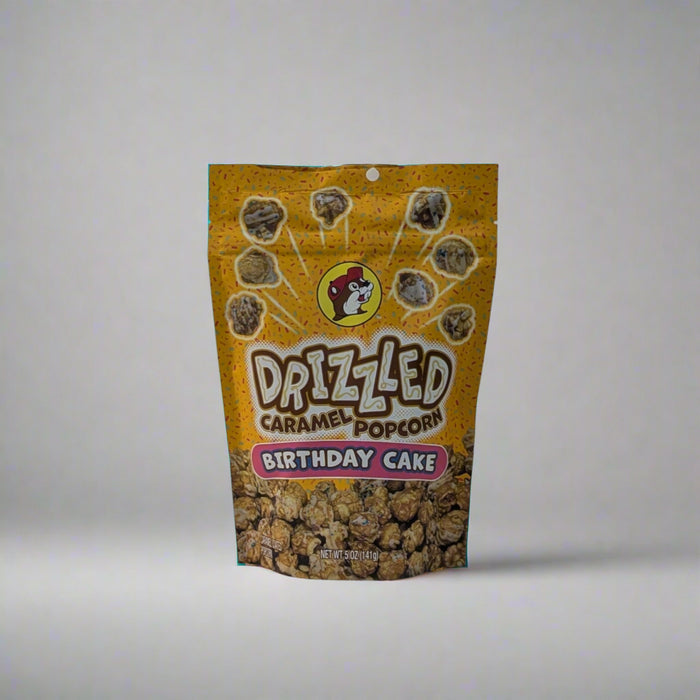 Two bags of Buc-ee’s Drizzled Caramel Popcorn, one in Black and White flavor and the other in Birthday Cake flavor, displayed on a white sheet backdrop.