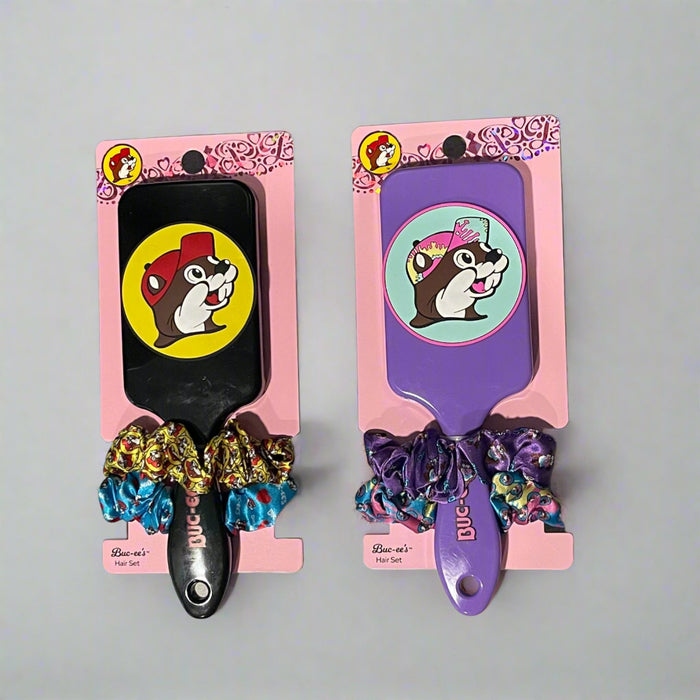 Two Buc-ee’s hair brush and scrunchie sets on display: one with a purple brush and heart scrunchies, the other with a black brush and colorful red-cap Buc-ee's theme scrunchies.