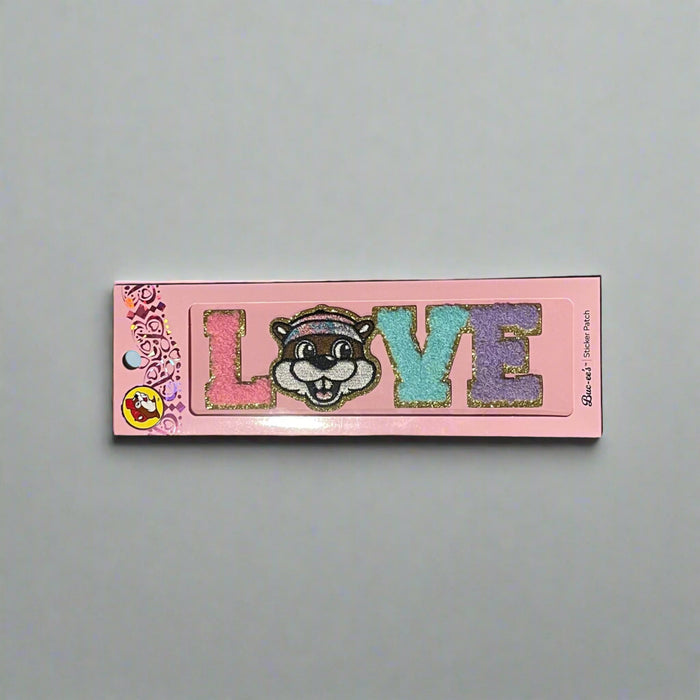 Two Buc-ee’s sticker patches shown on pink packaging: one spells “LOVE” with Buc-ee’s face as the “O,” and the other spells “PEACE” with Buc-ee in the center, both in colorful glitter letters.