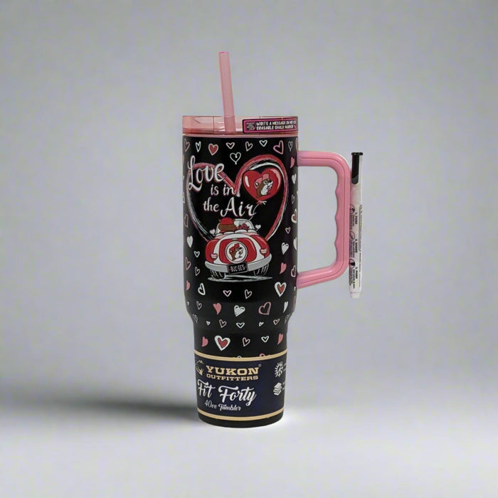 A black Valentine’s-themed tumbler featuring Buc-ee’s “Love is in the Air” design with hearts, a red car, and romantic details. The tumbler has a pink handle, reusable pink straw, and is crafted by Yukon Outfitters with a 40 oz capacity.