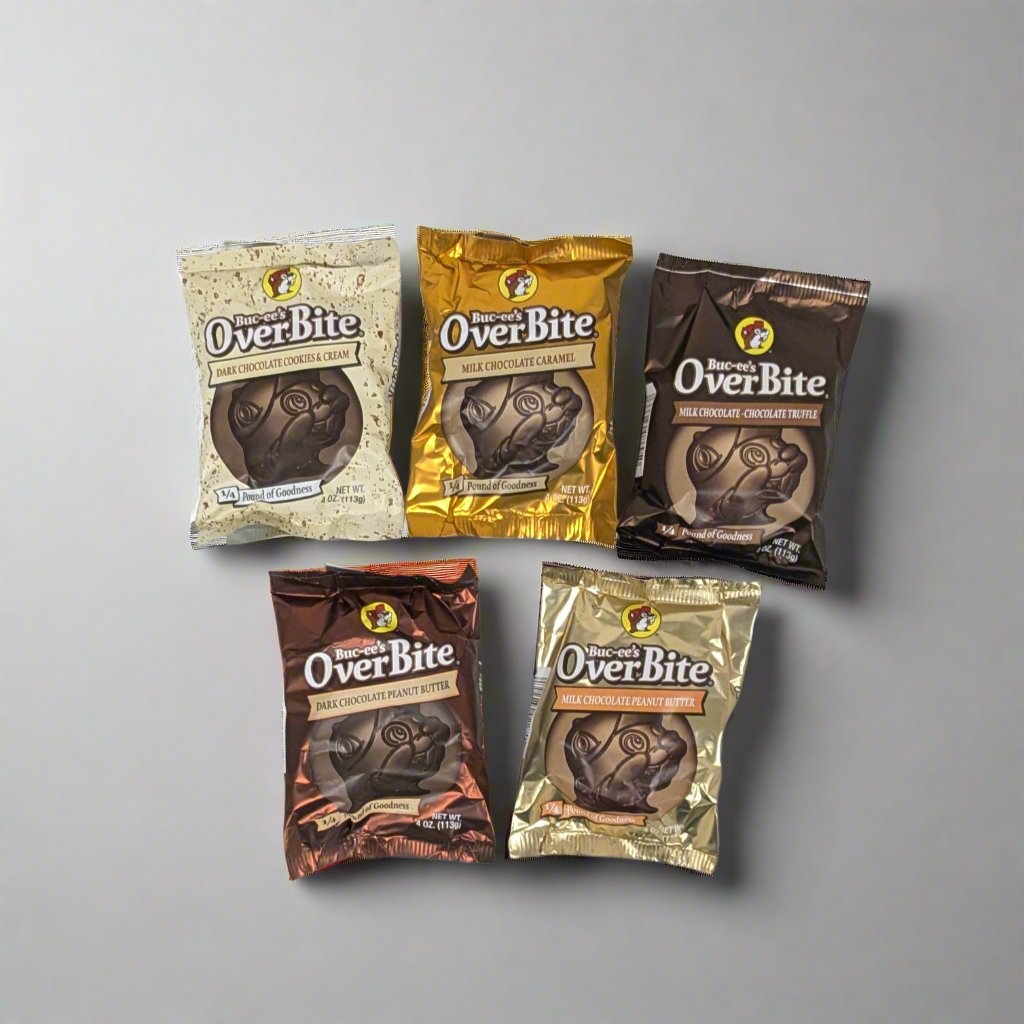 Five packages of "OverBite" chocolates are laid out on a white surface. The individually wrapped chocolates come in a variety of packaging colors, including brown, gold, cream, and bronze, each featuring an illustration of a beaver character wearing a hat. The arrangement suggests different flavors or varieties of the product. A chair with striped upholstery and a pair of sneakers worn by an individual standing on it are visible in the background.