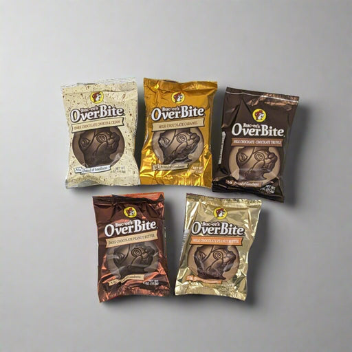 Five packages of "OverBite" chocolates are laid out on a white surface. The individually wrapped chocolates come in a variety of packaging colors, including brown, gold, cream, and bronze, each featuring an illustration of a beaver character wearing a hat. The arrangement suggests different flavors or varieties of the product. A chair with striped upholstery and a pair of sneakers worn by an individual standing on it are visible in the background.