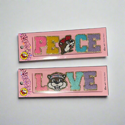Two Buc-ee’s sticker patches shown on pink packaging: one spells “LOVE” with Buc-ee’s face as the “O,” and the other spells “PEACE” with Buc-ee in the center, both in colorful glitter letters.