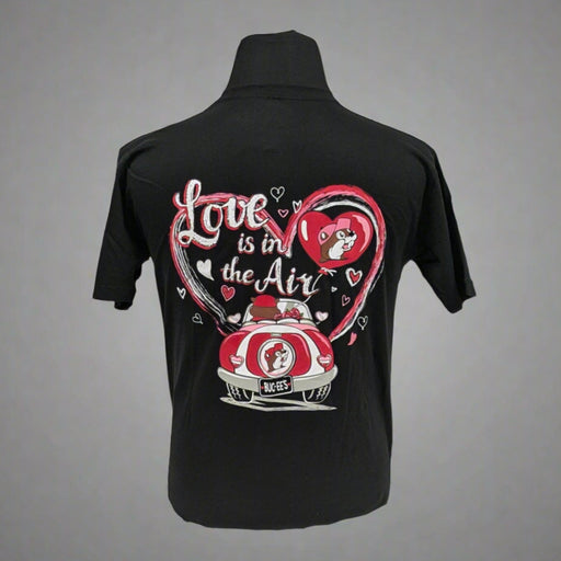 A black T-shirt featuring a colorful Valentine's Day-themed design on the back. The graphic includes two characters in a red car with a Buc-ee's logo on the license plate, surrounded by a large pink heart and smaller white hearts. The text reads "Love is in the Air" in a playful script font.