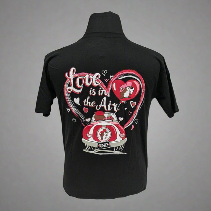 A black T-shirt featuring a colorful Valentine's Day-themed design on the back. The graphic includes two characters in a red car with a Buc-ee's logo on the license plate, surrounded by a large pink heart and smaller white hearts. The text reads "Love is in the Air" in a playful script font.