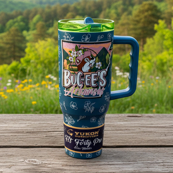 A blue handled tumbler with a green lid sits on a black tray in a store aisle. The tumbler features a colorful graphic of a mouse holding a picket sign that reads 'Bucee's Arkansas' and 'New York No.