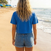 Person wearing a blue shirt and denim shorts standing on a beach with palm trees and ocean waves in the background.