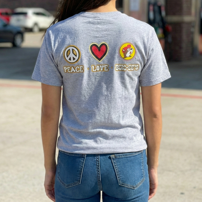 Person wearing a gray t-shirt with peace, love, and friendship patches in front of a gas station.
