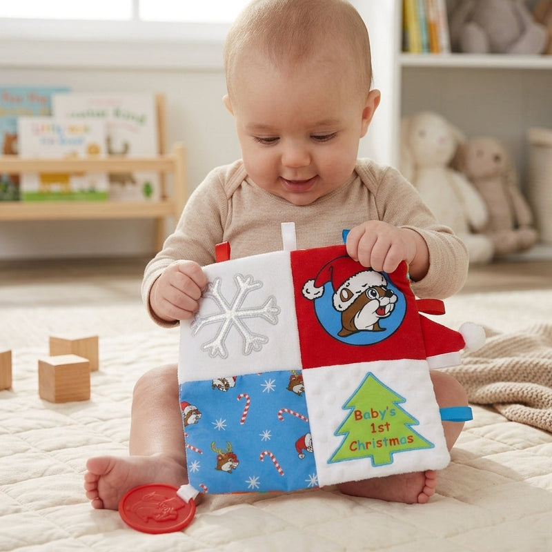 A baby sitting on a play mat holding a square Buc-ee's 'Baby’s 1st Christmas' crinkle toy featuring snowflake, Santa beaver, and candy cane designs. A red beaver-shaped teether is on the floor. In the background are shelves with toys and books.