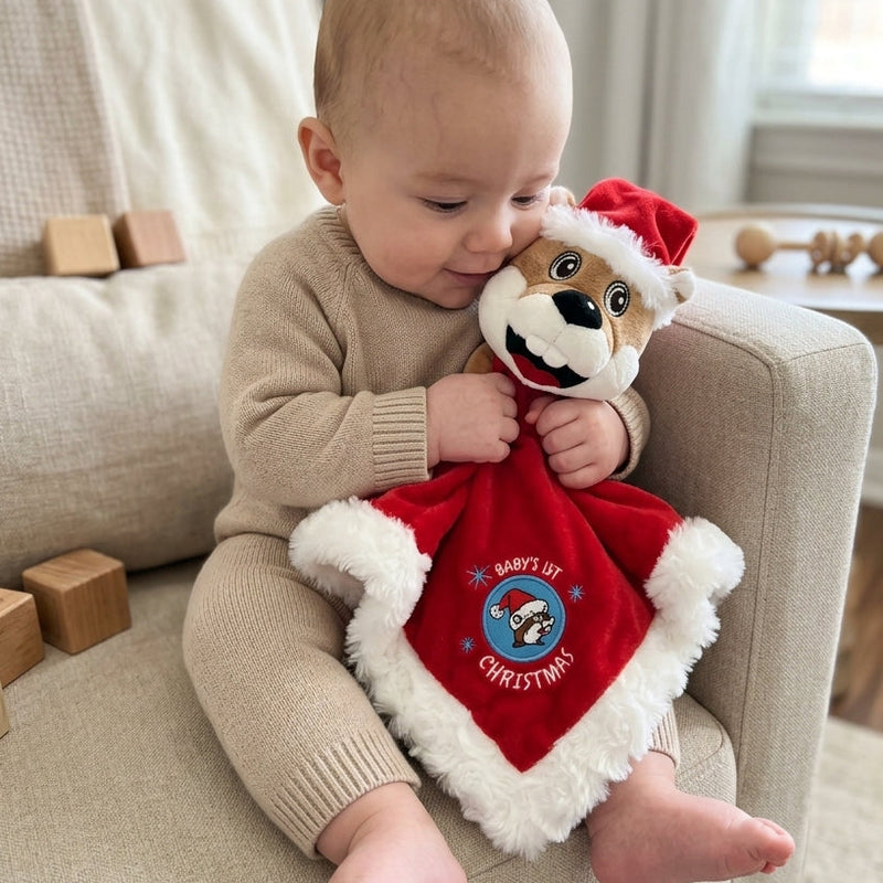 A close-up shot of a baby in a beige knitted onesie sitting in a light beige armchair, hugging a plush Buc-ee's Santa Beaver security blanket with red velvet fabric, white fur trim, and the embroidered text "BABY'S 1ST CHRISTMAS"