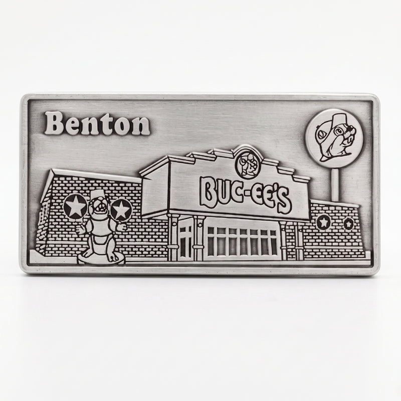 A rectangular silver-toned metal buckle featuring an embossed relief design of the fictional Benton BUC-EE's store and mascot, shown on a clean white background.