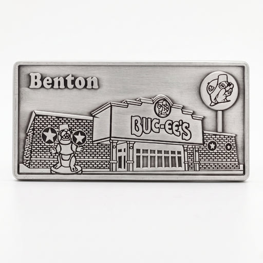 A rectangular silver-toned metal buckle featuring an embossed relief design of the fictional Benton BUC-EE's store and mascot, shown on a clean white background.