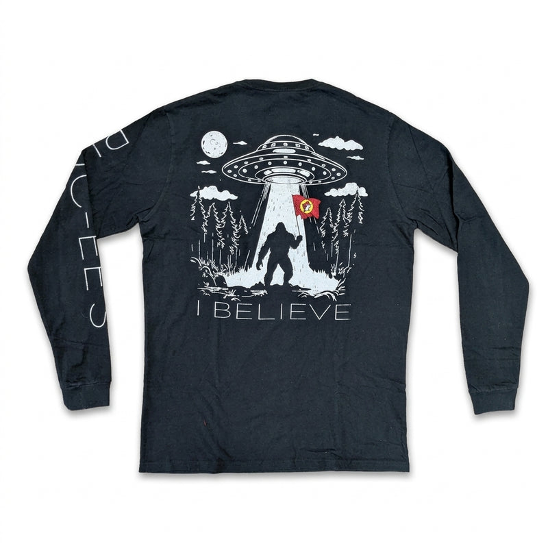 Back view of a black long-sleeve T-shirt featuring a UFO beaming up Bigfoot holding a red Buc-ee's flag in a pine forest above the text "I BELIEVE".