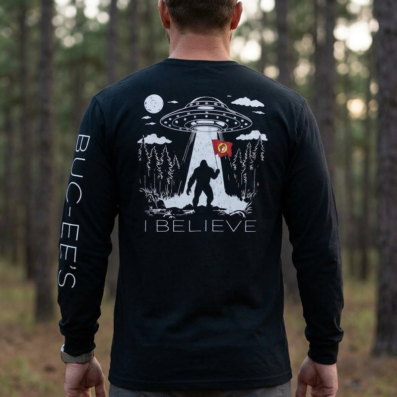 Back view of a man standing outdoors in a forest, wearing a black long-sleeve T-shirt featuring a detailed white graphics of Bigfoot beamed up by a UFO while holding a red Buc-ee's flag, with "I BELIEVE" printed below and "BUC-EE'S" down the left sleeve.