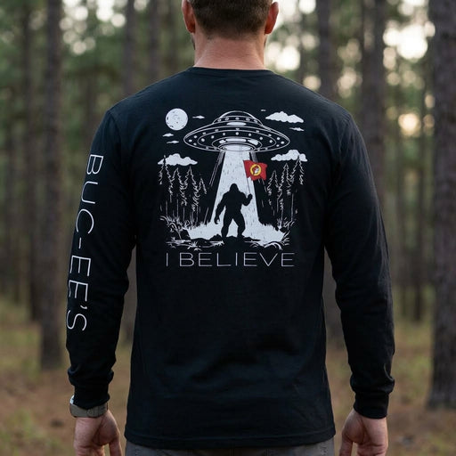 Back view of a man standing outdoors in a forest, wearing a black long-sleeve T-shirt featuring a detailed white graphics of Bigfoot beamed up by a UFO while holding a red Buc-ee's flag, with "I BELIEVE" printed below and "BUC-EE'S" down the left sleeve.