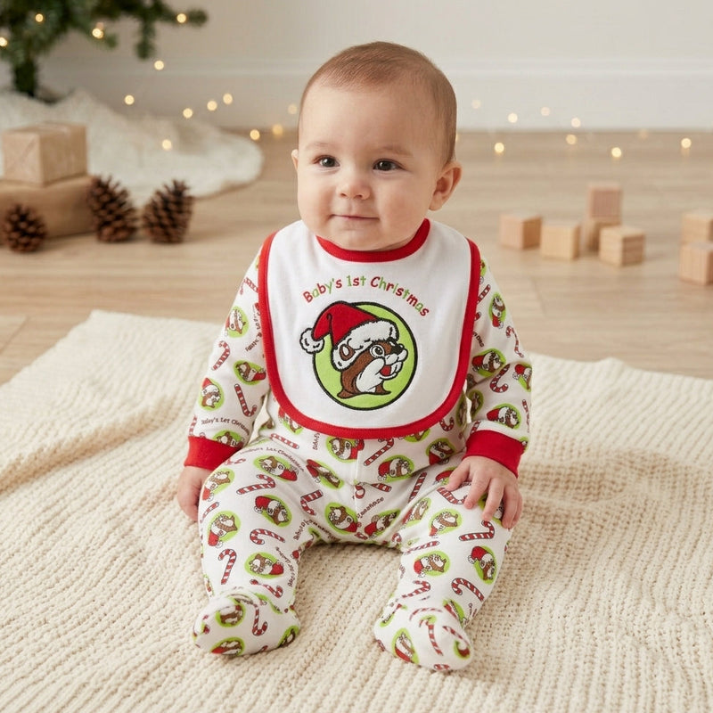 A close-up photograph features a baby sitting up on a textured white knitted blanket. The baby is wearing matching white and red-trimmed footie pajamas and a bib, both with an all-over pattern of red-and-white candy canes and the Buc-ee's beaver logo in a Santa hat, with the bib prominently displaying the text 'Baby's 1st Christmas' above a large beaver.