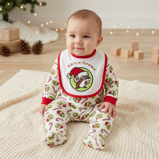 A close-up photograph features a baby sitting up on a textured white knitted blanket. The baby is wearing matching white and red-trimmed footie pajamas and a bib, both with an all-over pattern of red-and-white candy canes and the Buc-ee's beaver logo in a Santa hat, with the bib prominently displaying the text 'Baby's 1st Christmas' above a large beaver.