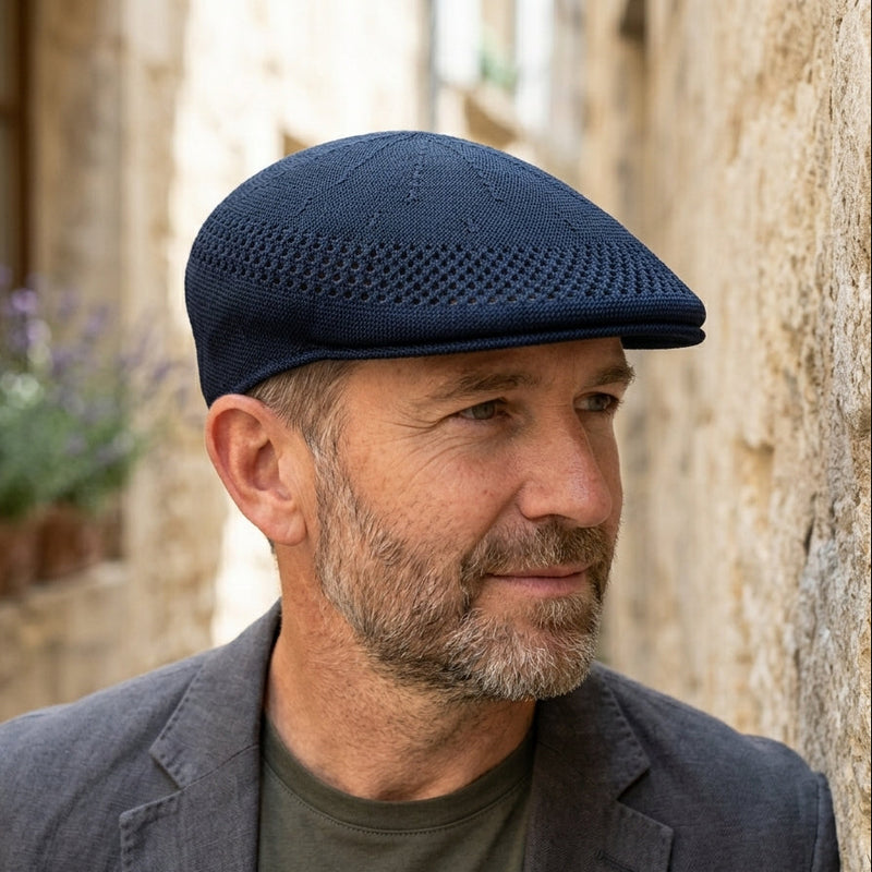 Close-up lifestyle portrait of a man with light stubble wearing a dark navy blue knit driver cap, looking off-camera against a soft-focus stone wall background.