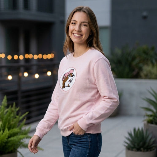 A woman standing outdoors smiling, wearing a light pink Buc-ee's adult sweatshirt with a circular chenille logo patch of the beaver mascot and blue jeans.