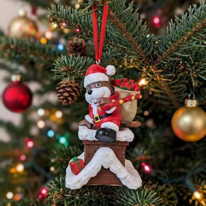 Close-up lifestyle image of the Buc-ee's Santa Beaver Christmas ornament hanging on a pine tree by its red ribbon. The beaver is dressed in a Santa suit on a snow-capped chimney, set against a softly blurred background of pine needles, pinecones, and colorful bokeh tree lights.