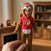 A hand holding a Barbie-sized fashion doll dressed in a Buc-ee’s beaver costume suit, standing in a cozy, warm-toned dollhouse living room with a wooden fireplace and bookshelf in the background.