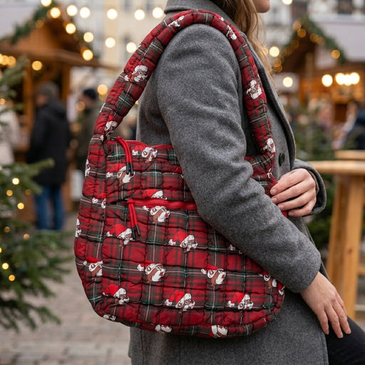 A person wearing a textured grey coat carries a Buc-ee's Christmas Red Puffy Tote Bag over their right shoulder. The large bag features a red and green tartan plaid pattern with a repeating print of a cartoon Buc-ee beaver wearing a Santa hat. The person's right hand is visible. The background is a blurred view of an outdoor market with warm string lights and pine trees at dusk.