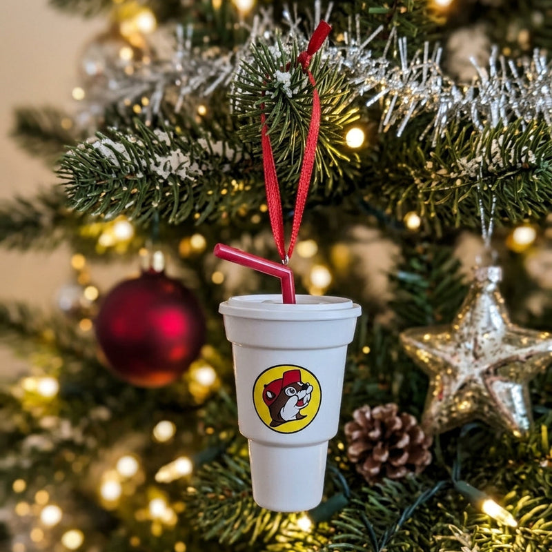 Close-up of a Buc-ee’s white cup Christmas ornament with a red straw and beaver logo hanging from a pine tree branch surrounded by warm holiday lights and silver tinsel.