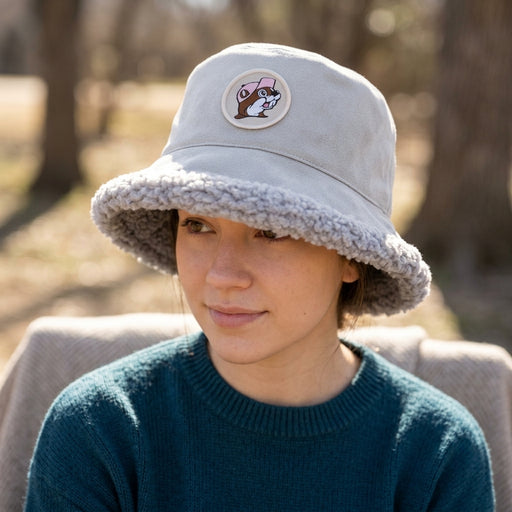 Close-up lifestyle portrait of a woman wearing a Buc-ee's light gray faux-suede bucket hat with plush sherpa brim trim and a circular Buc-ee's beaver logo patch. She is wearing a dark teal sweater in a blurred, natural outdoor setting.
