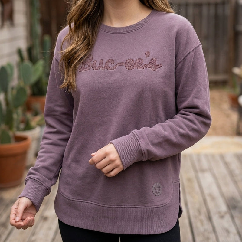 A smiling woman wearing a mauve-purple crewneck sweatshirt with 'Buc-ee's' embroidered across the chest, standing on a wooden deck with string lights and potted cacti in the background.