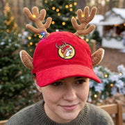 Close-up lifestyle shot of a woman outdoors during winter, wearing a red corduroy Buc-ee's reindeer baseball hat. The hat features plush antlers wrapped in lighted string lights, plush ears, and an embroidered holiday beaver logo.