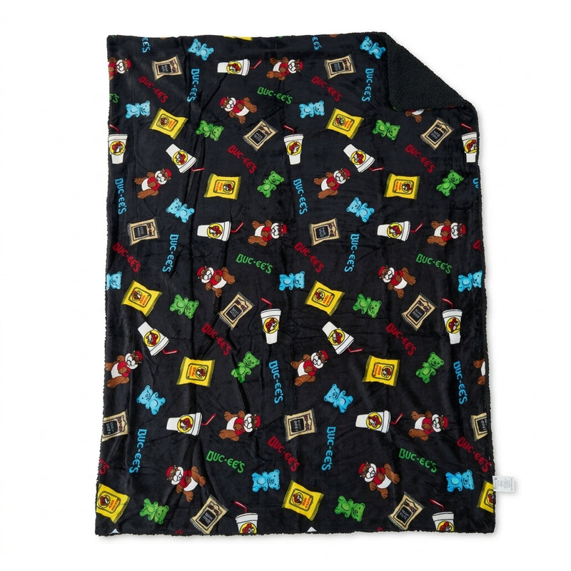 Black Buc-ee's sherpa-lined throw blanket laid flat, featuring a colorful print of Beaver Nuggets, drinks, gummy bears, beef jerky, and Buc-ee's logos.