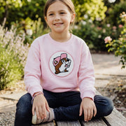 Young girl wearing the light pink Buc-ee's youth crewneck sweatshirt featuring a textured chenille beaver logo patch, sitting outdoors on a wooden bench.