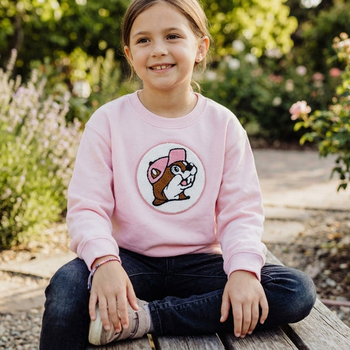 Young girl wearing the light pink Buc-ee's youth crewneck sweatshirt featuring a textured chenille beaver logo patch, sitting outdoors on a wooden bench.