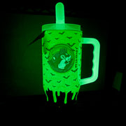 A glow-in-the-dark green tumbler cup with a handle, featuring a cartoon skull graphic and dripping slime design, shown glowing in the dark.