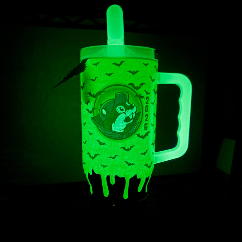 A glow-in-the-dark green tumbler cup with a handle, featuring a cartoon skull graphic and dripping slime design, shown glowing in the dark.