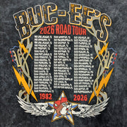 An up close photo of the graphic on the back of a Buc-ee's 2026 Tour Tee, a black mineral-wash t-shirt featuring a large, concert-style graphic titled "BUC-EE'S 2026 ROAD TOUR." The center contains a comprehensive, three-column list of Buc-ee’s locations and their opening years, starting from 1982 in Lake Jackson, TX, through 2026. The list is framed by golden lightning bolts and electric guitars, with a small Buc-ee's beaver logo set inside a star with white wings at the bottom.