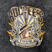 Up close picture of the graphic on the front of the 2026 Tour Tee, a black mineral-wash t-shirt featuring a large, vibrant rock-and-roll graphic. In the center, the Buc-ee’s beaver wears a stylish black fedora, surrounded by electric guitars, radiant white wings, golden lightning bolts, and vintage-style speakers. A ribbon banner at the bottom reads "ROAD TOUR 2026" and "ROCKIN' SINCE '82," all set against a blue and yellow sunburst background.