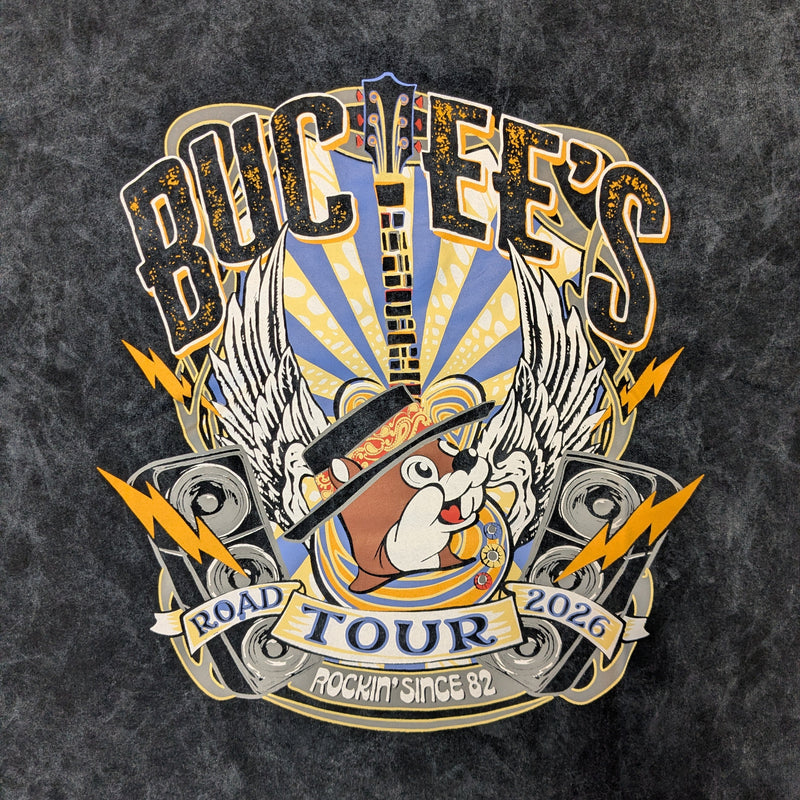 Up close picture of the graphic on the front of the 2026 Tour Tee, a black mineral-wash t-shirt featuring a large, vibrant rock-and-roll graphic. In the center, the Buc-ee’s beaver wears a stylish black fedora, surrounded by electric guitars, radiant white wings, golden lightning bolts, and vintage-style speakers. A ribbon banner at the bottom reads "ROAD TOUR 2026" and "ROCKIN' SINCE '82," all set against a blue and yellow sunburst background.