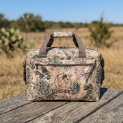 A front view of the Buc-ee's GameGuard 24-Can Cooler bag resting on a weathered wooden picnic table. The cooler features a realistic brush country camouflage print with cactus designs and an embroidered Buc-ee's logo on the front pocket. The background shows a soft-focus view of a sunny, open Texas field under a clear blue sky.