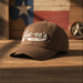 Close-up of a brown Buc-ee’s “Est. 1982” baseball cap resting on a rustic wooden surface with warm sunlight. The hat’s white embroidered logo is crisp and centered, with a blurred Texas flag and star décor in the background, creating a cozy Texas atmosphere.