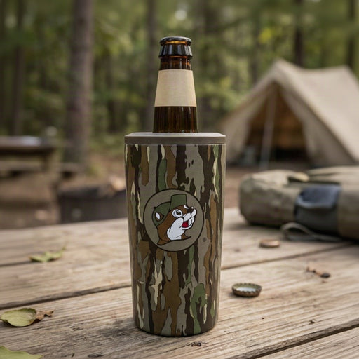 A close-up lifestyle shot of a Buc-ee's camouflage 4-in-1 drink cooler sitting on an outdoor wooden picnic table. The cooler features the Buc-ee's beaver mascot wearing a camo cap in a circular logo. Inside the cooler is a 12oz glass bottle with a plain kraft neck label. In the background, a tent is pitched among surrounding trees in a forest campsite setting.