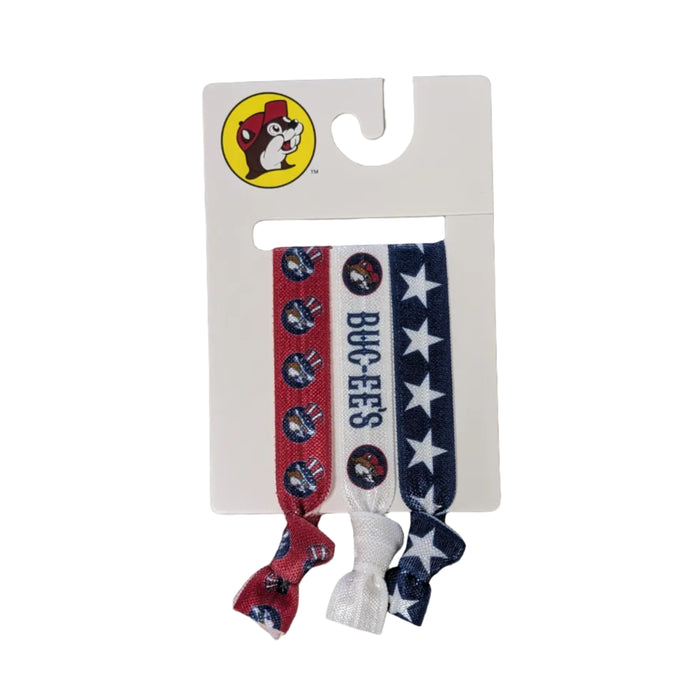 A set of three Buc-ee's branded elastic ribbon hair ties on a white cardboard display card. The first tie is red with a repeating pattern of Buc-ee the beaver wearing a patriotic Uncle Sam hat. The middle tie is white with the word "BUC-EE'S" in blue and two beaver logos. The third tie is navy blue with a repeating white star pattern. Each tie is knotted at the bottom.