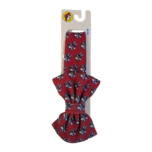 A fabric-covered headband with a large matching red bow. The fabric features a repeating print of the Buc-ee's beaver wearing an American flag top hat. It is displayed on an official white Buc-ee's retail card.
