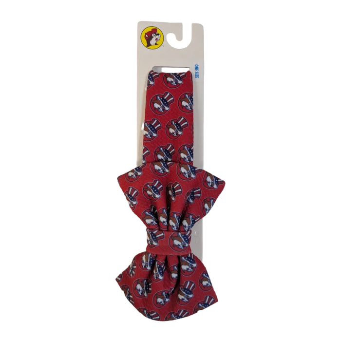A fabric-covered headband with a large matching red bow. The fabric features a repeating print of the Buc-ee's beaver wearing an American flag top hat. It is displayed on an official white Buc-ee's retail card.