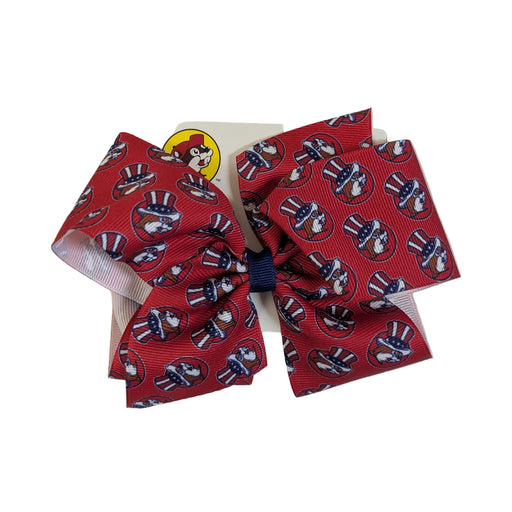 A close-up of a Buc-ee's patriotic hair bow made of red grosgrain ribbon. The ribbon features a repeating print of the Buc-ee's beaver mascot wearing a red, white, and blue American flag top hat. The bow is tied with a navy blue center knot and is attached to an official white Buc-ee's retail hanging card. Displayed on a clean white background.