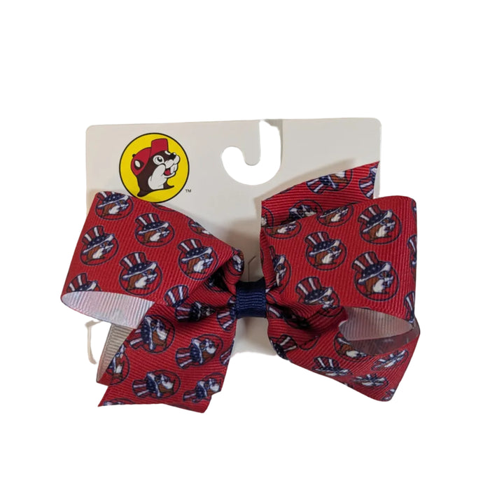 A close-up of a Buc-ee's patriotic hair bow made of red grosgrain ribbon. The ribbon features a repeating print of the Buc-ee's beaver mascot wearing a red, white, and blue American flag top hat. The bow is tied with a navy blue center knot and is attached to an official white Buc-ee's retail hanging card. Displayed on a clean white background.