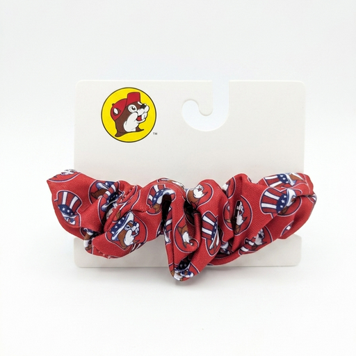 A red fabric hair scrunchie featuring a repeating print of the Buc-ee's beaver mascot wearing a patriotic American flag top hat. It is displayed on a white Buc-ee's retail hanging card.